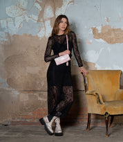 In a room with peeling walls and a brick floor, a woman in a black lace dress leans against a worn-out chair, holding the Cadence clutch by Bed Stu.