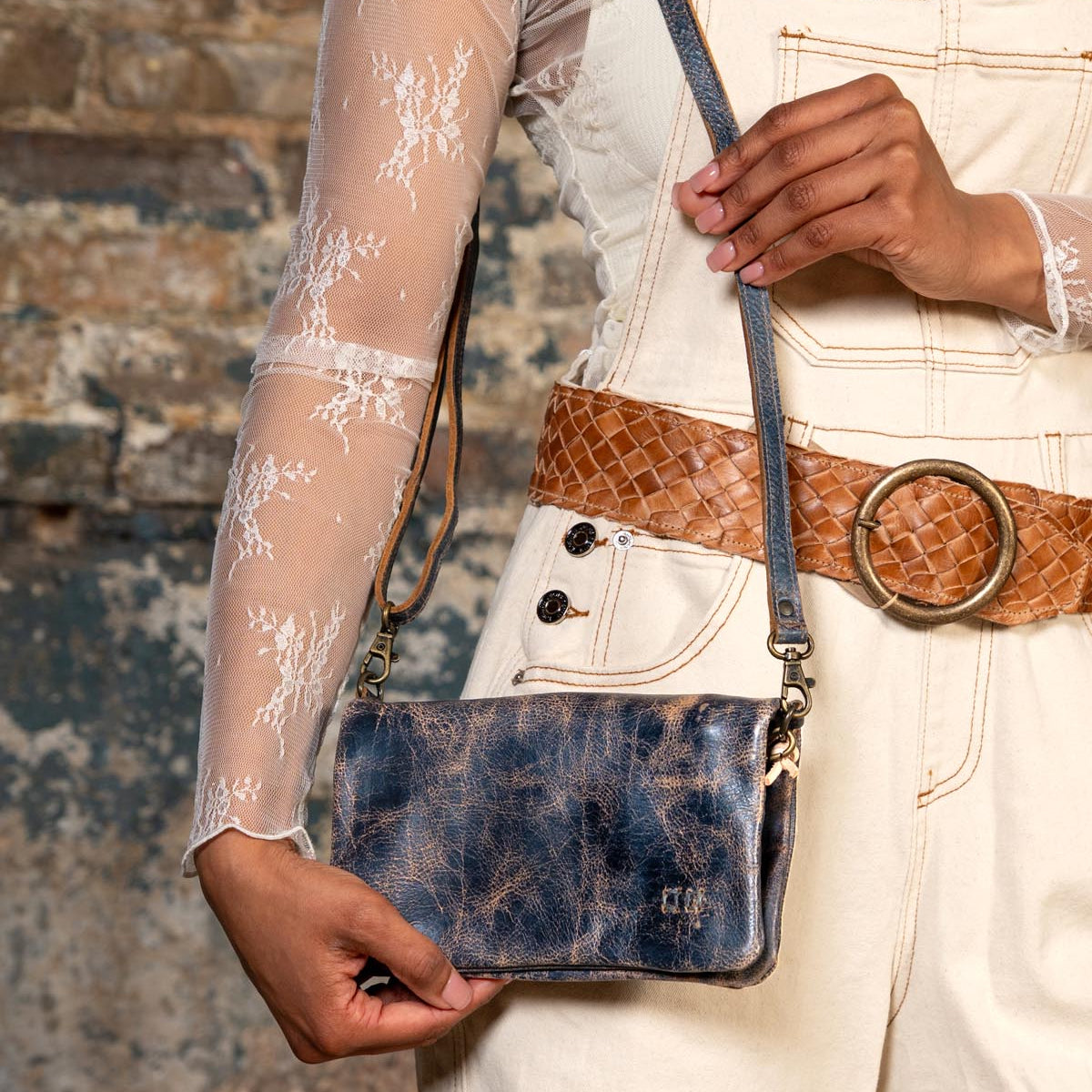 A person holds the Bed Stu Cadence distressed blue leather clutch against a textured brick wall, styled with a sheer lace top, cream overalls, and a brown woven belt.