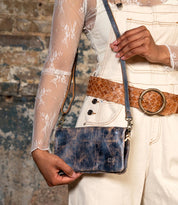 A person holds the Bed Stu Cadence distressed blue leather clutch against a textured brick wall, styled with a sheer lace top, cream overalls, and a brown woven belt.