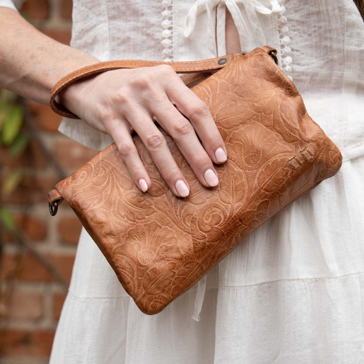 Wearing a white dress, someone holds the Bed Stu Cadence EM embossed leather crossbody clutch with a floral pattern while standing in front of a brick wall.