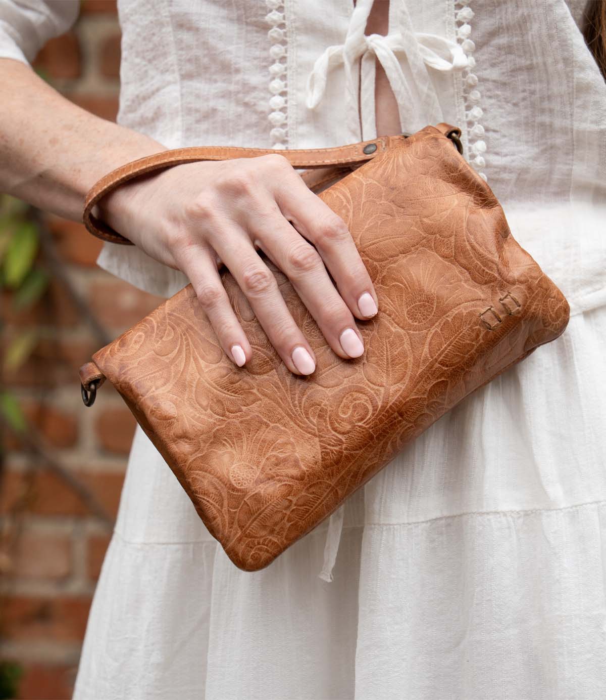 Wearing a white dress, someone holds the Bed Stu Cadence EM embossed leather crossbody clutch with a floral pattern while standing in front of a brick wall.