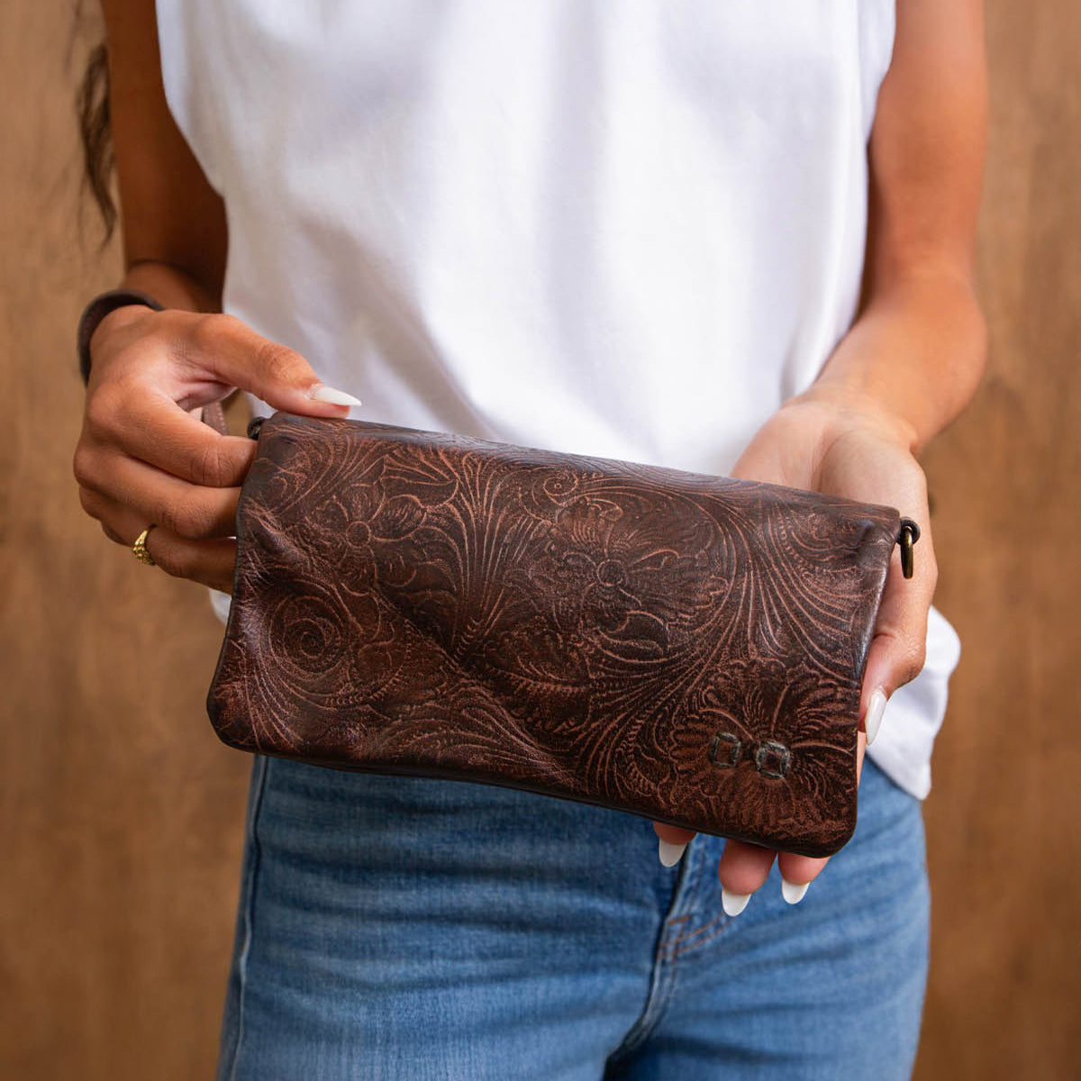 A person wearing a white sleeveless top and blue jeans holds the Cadence EM, a brown embossed leather floral clutch purse by Bed Stu.