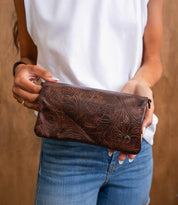 A person wearing a white sleeveless top and blue jeans holds the Cadence EM, a brown embossed leather floral clutch purse by Bed Stu.