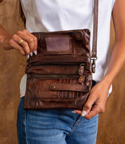 A person wearing a white shirt and blue jeans holds the Bed Stu Cadence EM brown leather crossbody bag with multiple compartments and a visible zipper, showcasing this stylish accessory.