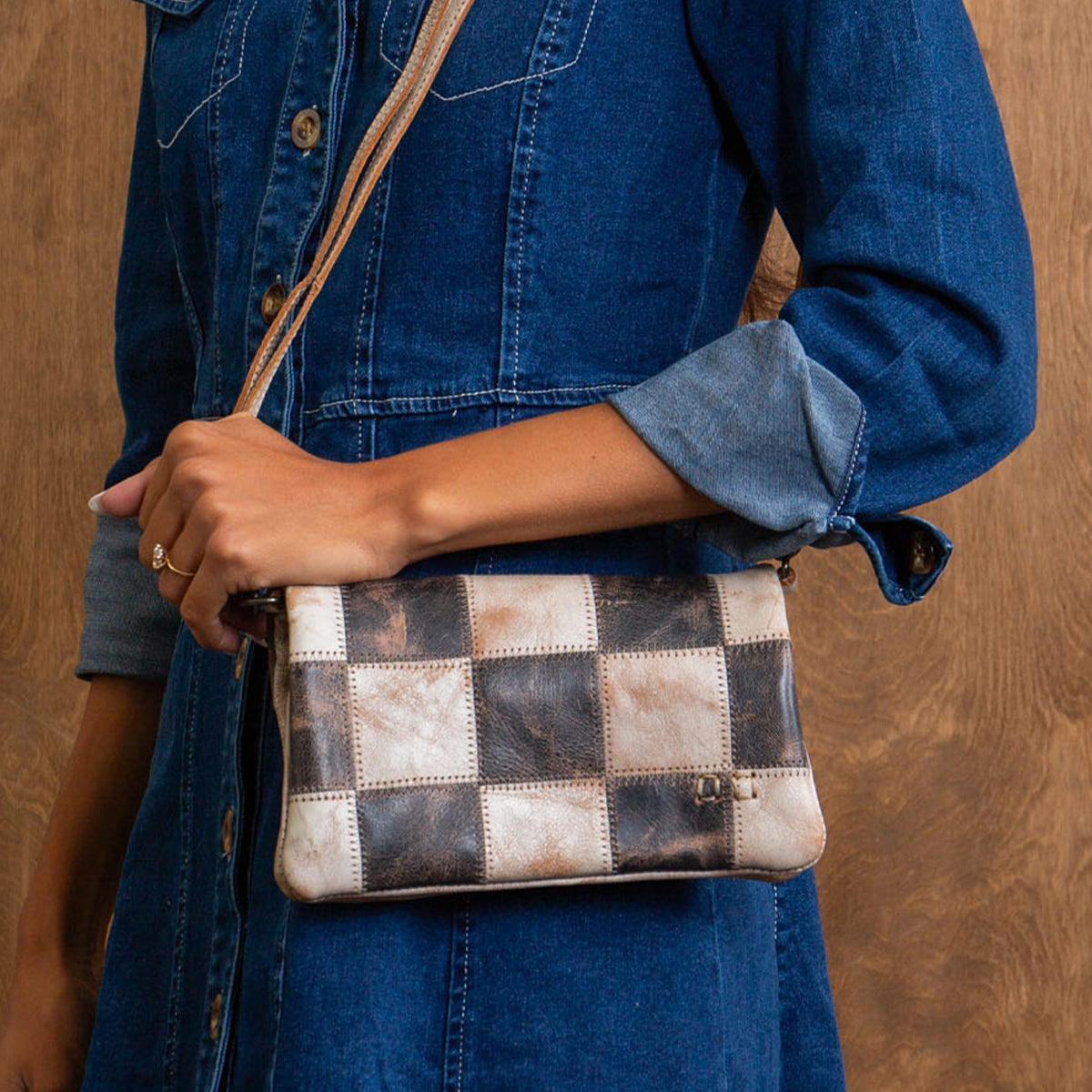 A person in a denim dress carries the Bed Stu Cadence PW—a small patchwork clutch styled as a leather crossbody with checkered pattern, ideal as a chic travel wallet.