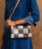 A person in a denim dress carries the Bed Stu Cadence PW—a small patchwork clutch styled as a leather crossbody with checkered pattern, ideal as a chic travel wallet.