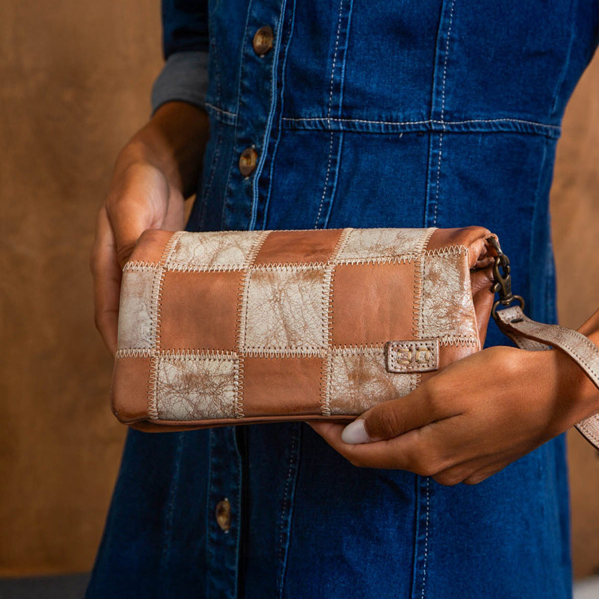A person in a blue denim dress holds the Bed Stu Cadence PW, a handcrafted clutch with brown and beige patchwork detailing, in one hand.