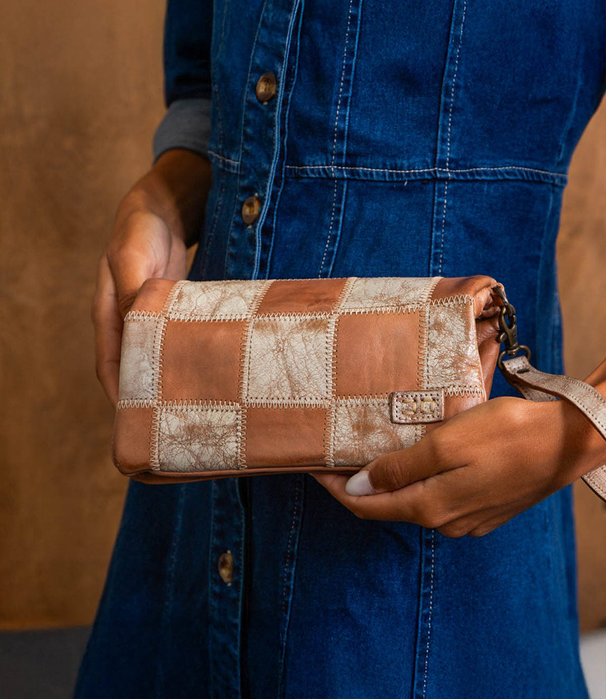 A person in a blue denim dress holds the Bed Stu Cadence PW, a handcrafted clutch with brown and beige patchwork detailing, in one hand.