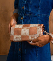 A person in a blue denim dress holds the Bed Stu Cadence PW, a handcrafted clutch with brown and beige patchwork detailing, in one hand.