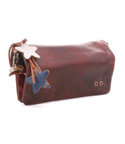 The Cadence by Bed Stu is a brown vegetable-tanned leather clutch with two star-shaped leather charms—one white, one blue—dangling from a strap attached to the zipper.