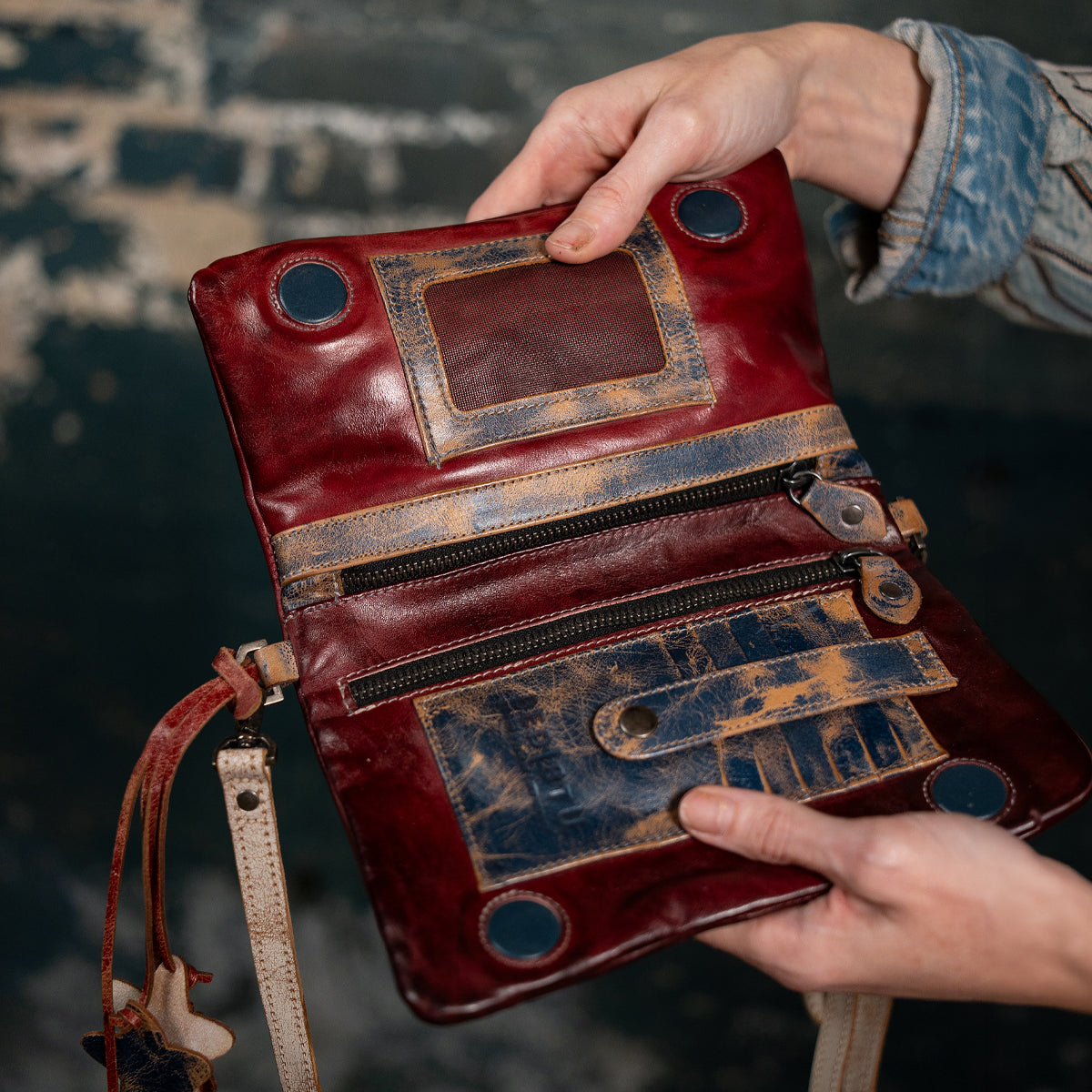 Someone holds the Bed Stu Cadence, an open crossbody clutch crafted from vegetable-tanned leather with zippers, pockets, and distressed details, set against a rustic dark background.