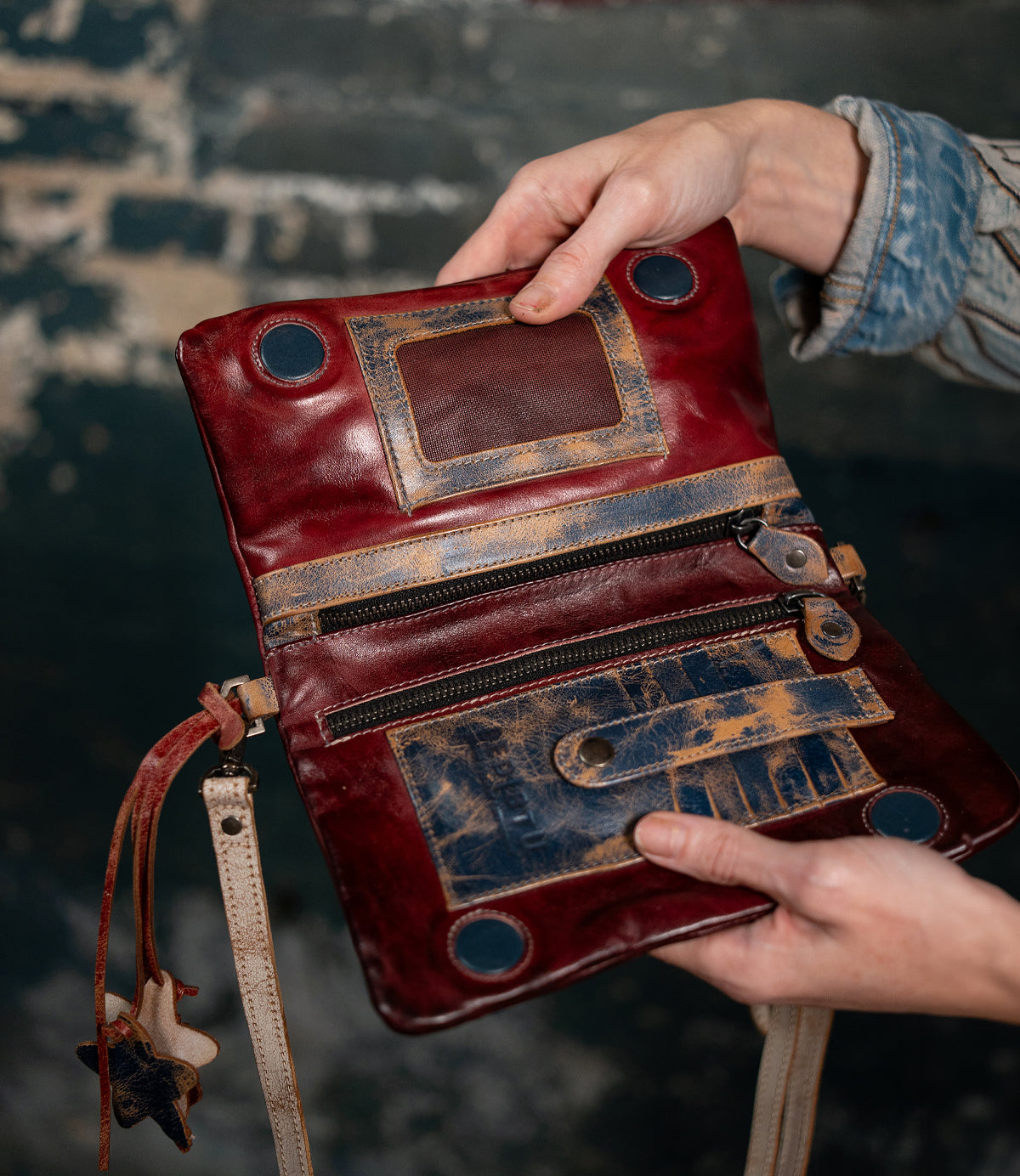 Someone holds the Bed Stu Cadence, an open crossbody clutch crafted from vegetable-tanned leather with zippers, pockets, and distressed details, set against a rustic dark background.
