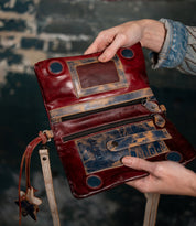 Someone holds the Bed Stu Cadence, an open crossbody clutch crafted from vegetable-tanned leather with zippers, pockets, and distressed details, set against a rustic dark background.