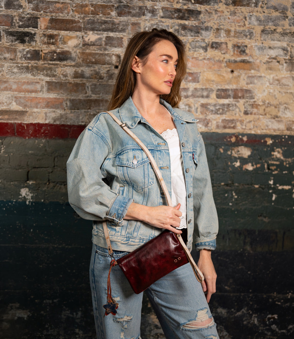 Woman in a denim jacket and ripped jeans poses by a brick wall, holding the Bed Stu Cadence—a small burgundy crossbody clutch made from vegetable-tanned leather.