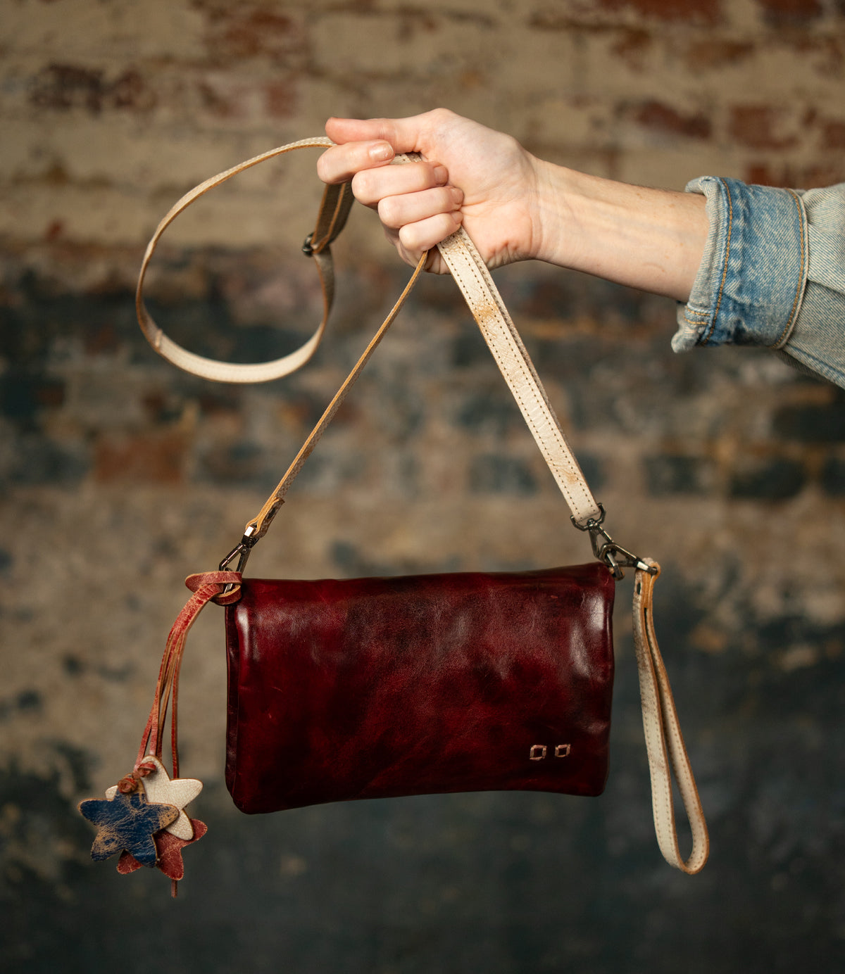 A hand in a denim sleeve holds the Bed Stu Cadence, a small, dark red vegetable-tanned leather crossbody clutch with a star-shaped keychain, set against a textured brick wall background.