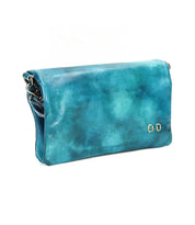The Cadence by Bed Stu is a turquoise crossbody clutch made from vegetable-tanned leather, featuring a flap closure and two small gold rectangular accents on the front.