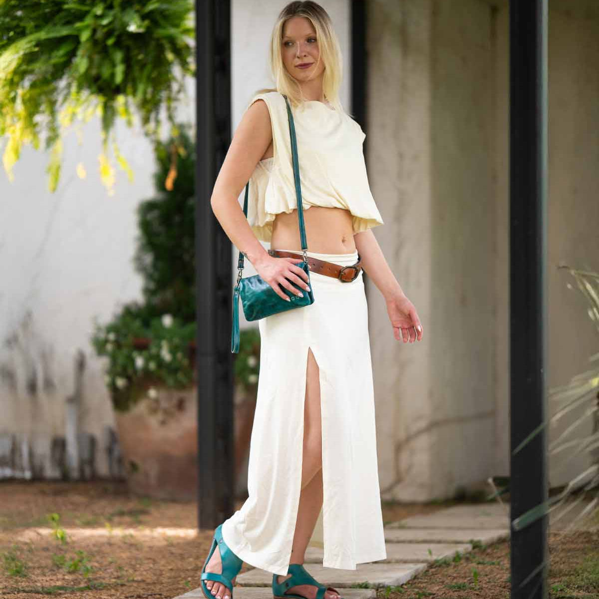 A person stands outdoors wearing a sleeveless cream crop top and matching skirt, brown belt, teal sandals and shoulder bag, and carrying the Bed Stu Cadence vegetable-tanned leather 3-in-1 wallet.