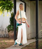 A person stands outdoors wearing a sleeveless cream crop top and matching skirt, brown belt, teal sandals and shoulder bag, and carrying the Bed Stu Cadence vegetable-tanned leather 3-in-1 wallet.