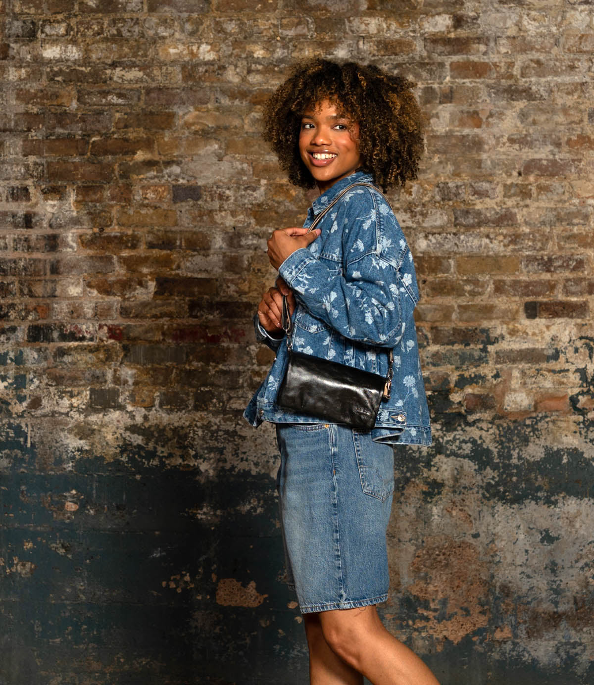 A person in a denim jacket and skirt stands sideways against a weathered brick wall, smiling while holding the Bed Stu Cadence black crossbody purse.