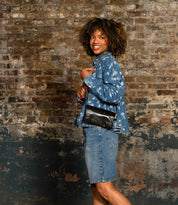 A person in a denim jacket and skirt stands sideways against a weathered brick wall, smiling while holding the Bed Stu Cadence black crossbody purse.