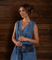 A woman stands in front of a wooden wall, wearing a sleeveless blue denim dress and holding the Bed Stu Cadence crossbody purse in dark navy leather.