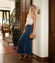 A woman stands indoors near a staircase, wearing a white strapless top, high-waisted wide-leg jeans, brown platform heels, and holding the Bed Stu Cadence tan vegetable-tanned leather crossbody clutch.