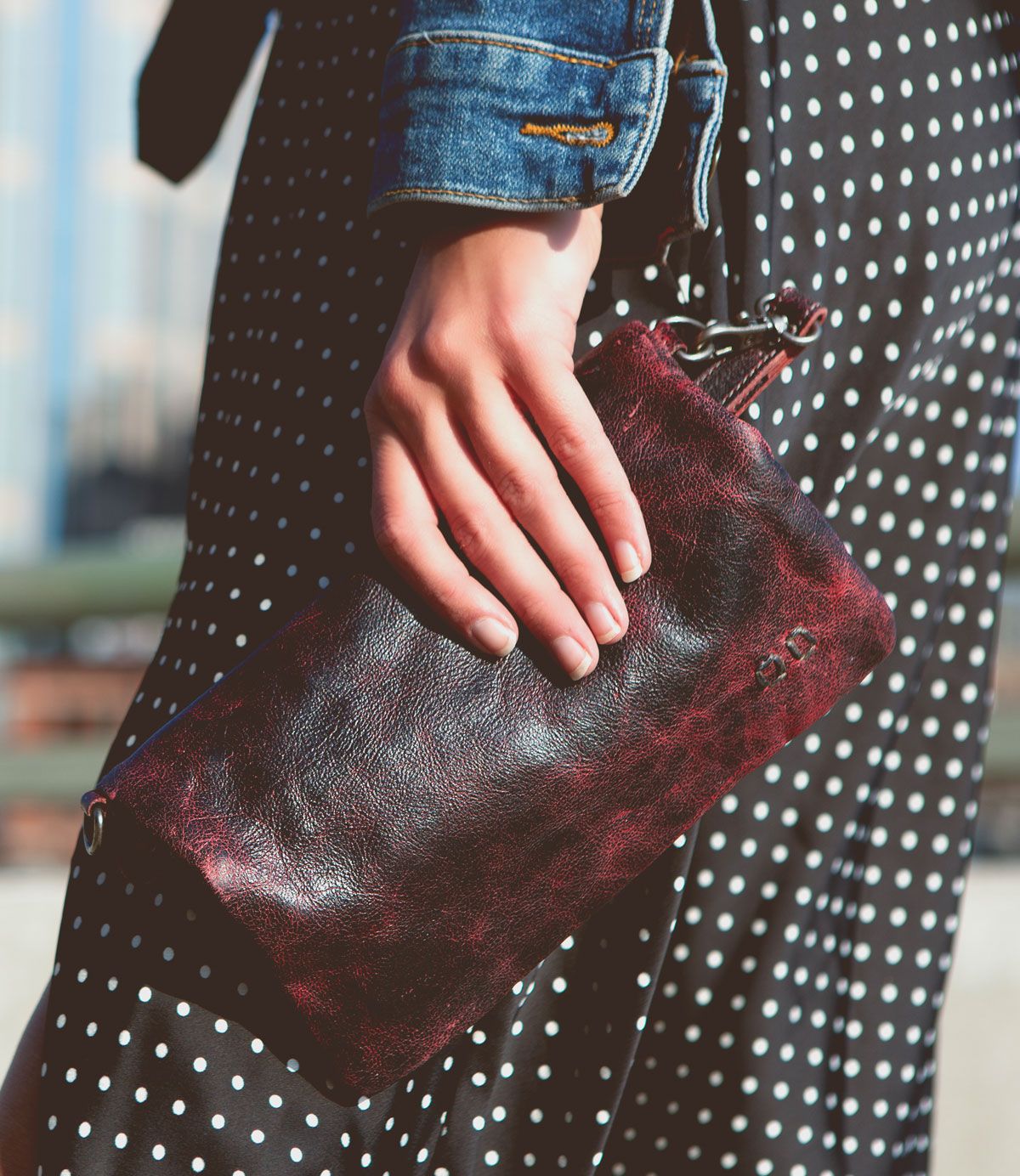 A close-up of a person’s hand holding the Cadence clutch bag in dark red leather by Bed Stu. The person is wearing a polka-dot dress and a denim jacket, stylishly pulling the look together.