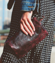 A close-up of a person’s hand holding the Cadence clutch bag in dark red leather by Bed Stu. The person is wearing a polka-dot dress and a denim jacket, stylishly pulling the look together.