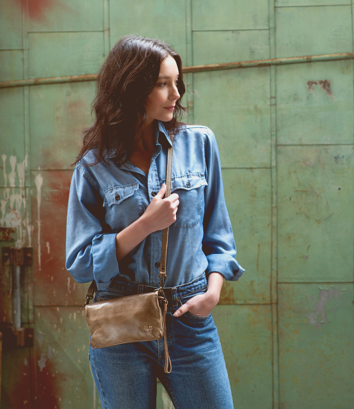 A woman wearing a denim shirt and jeans standing in front of a green wall with her Bed Stu Cadence crossbody bag.