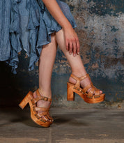 A person in a blue ruffled dress and Bed Stu Caitlin II sandals with a wooden heel and woven leather upper stands on a stone floor against a textured wall.