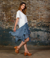 A woman stands sideways by a brick wall, wearing a white t-shirt, blue ruffled skirt, and Bed Stu Caitlin II platform sandals with a woven leather upper and stylish wooden heel.