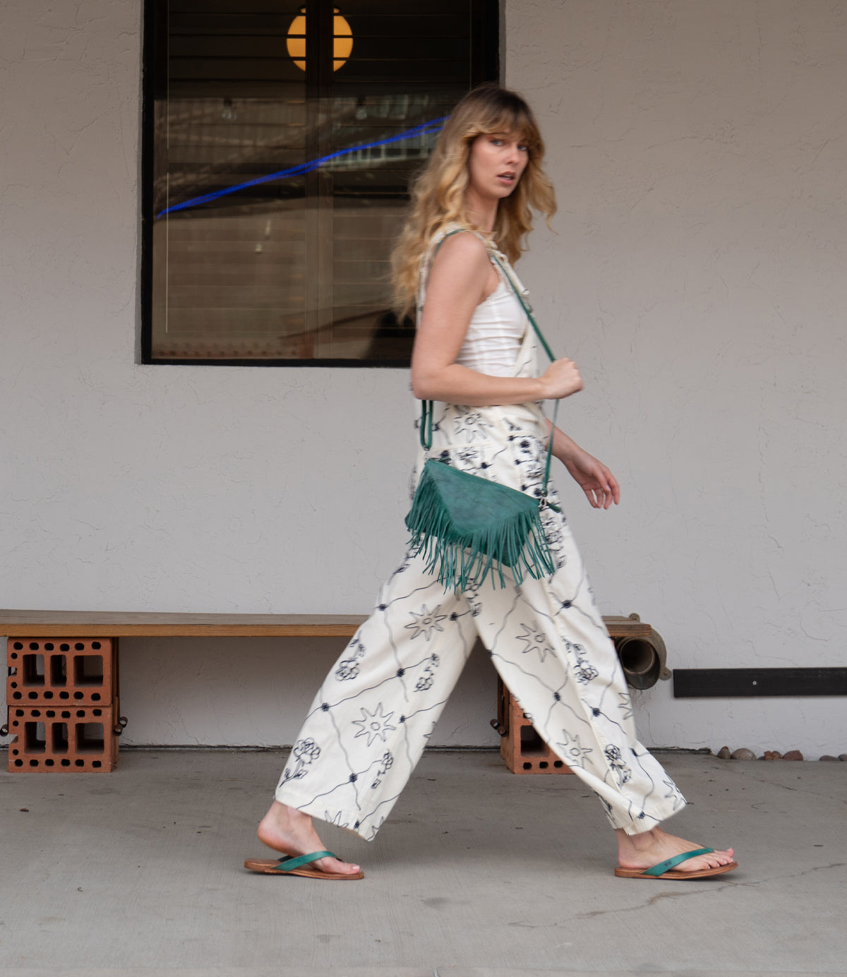 A woman with long hair walks past a beige wall and window, wearing a white patterned outfit, green fringe bag, and comfortable Calma sandals by Bed Stu.