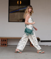 A woman with long hair walks past a beige wall and window, wearing a white patterned outfit, green fringe bag, and comfortable Calma sandals by Bed Stu.