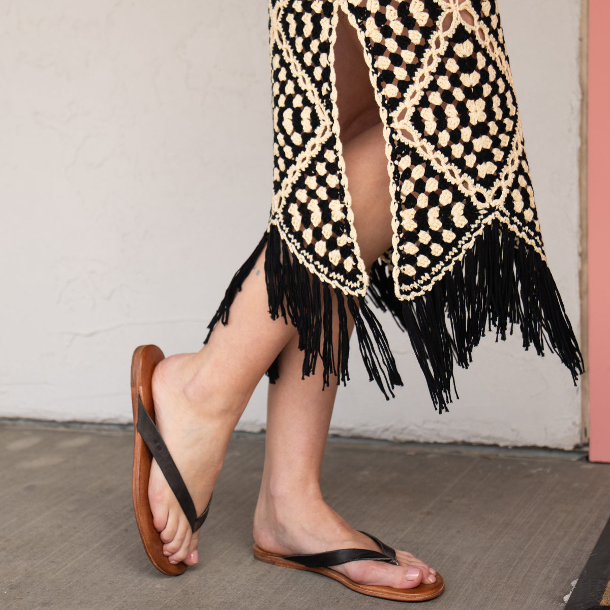 A person in a black and cream crochet skirt with fringe stands on a concrete floor, wearing Bed Stu Calma relaxing sandals.