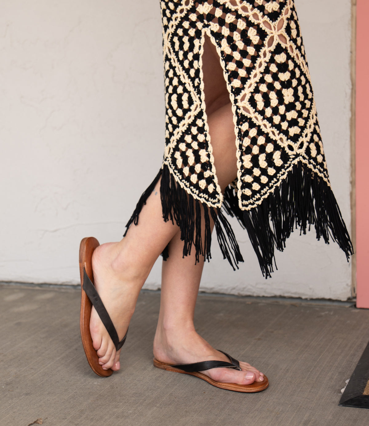 A person in a black and cream crochet skirt with fringe stands on a concrete floor, wearing Bed Stu Calma relaxing sandals.
