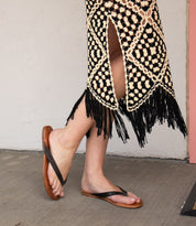 A person in a black and cream crochet skirt with fringe stands on a concrete floor, wearing Bed Stu Calma relaxing sandals.