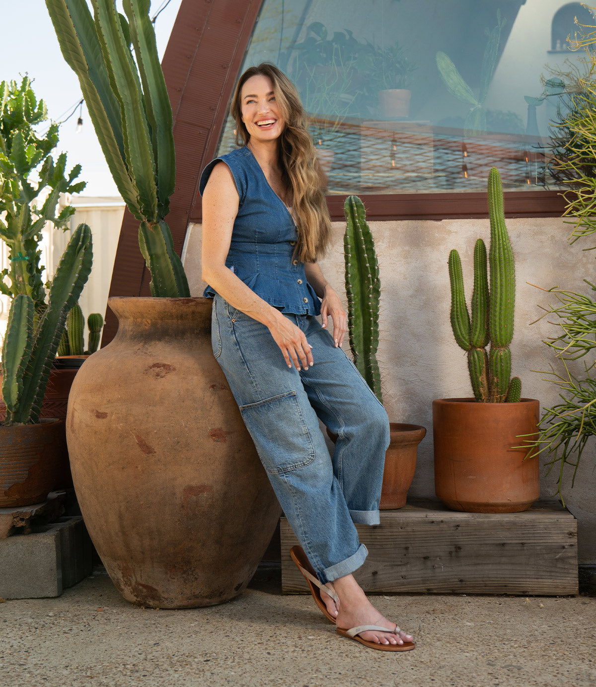 A woman in a blue top and jeans stands smiling outdoors by large clay pots and cacti, wearing Bed Stu's Calma lightweight, comfortable sandals.