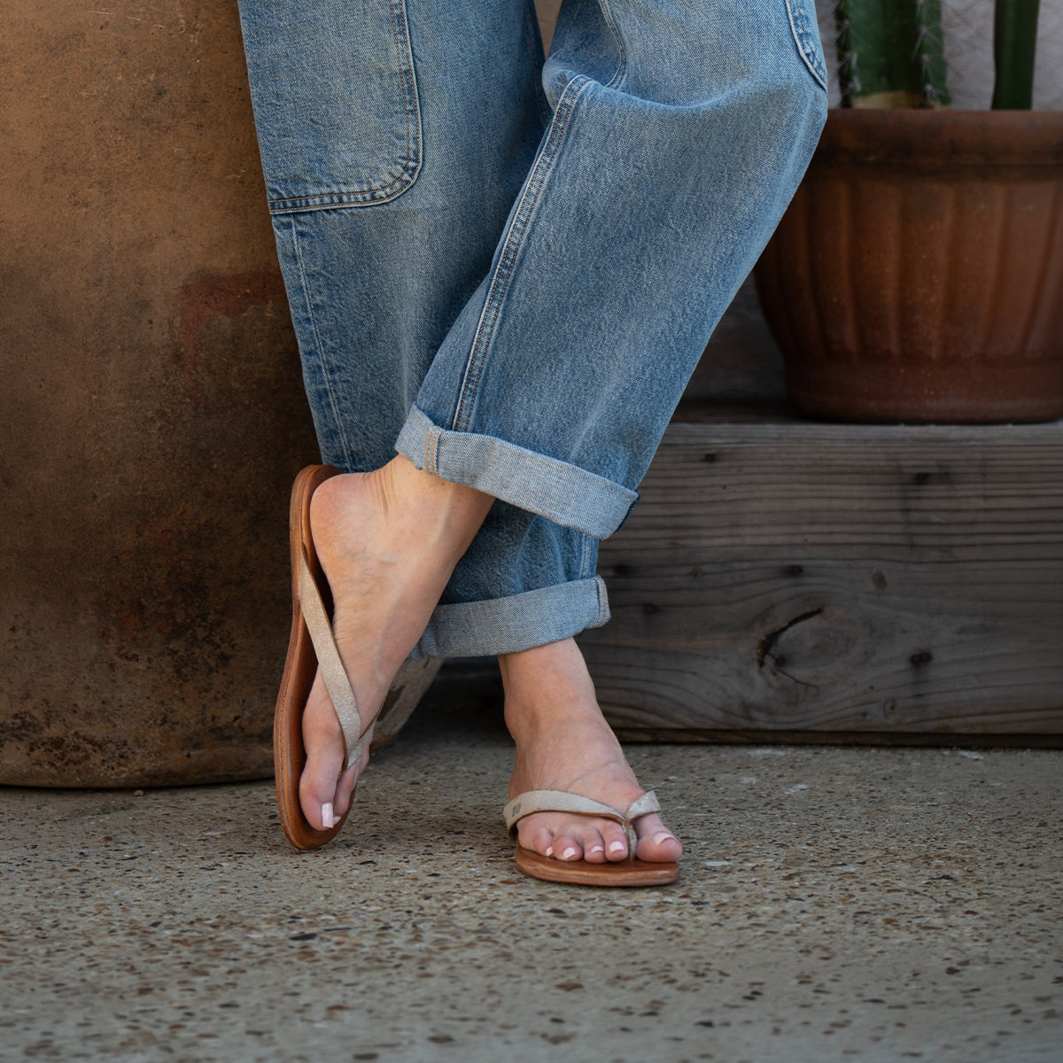 A person wearing rolled-up blue jeans and Bed Stu's Calma sandals stands on a textured concrete surface next to a potted cactus.
