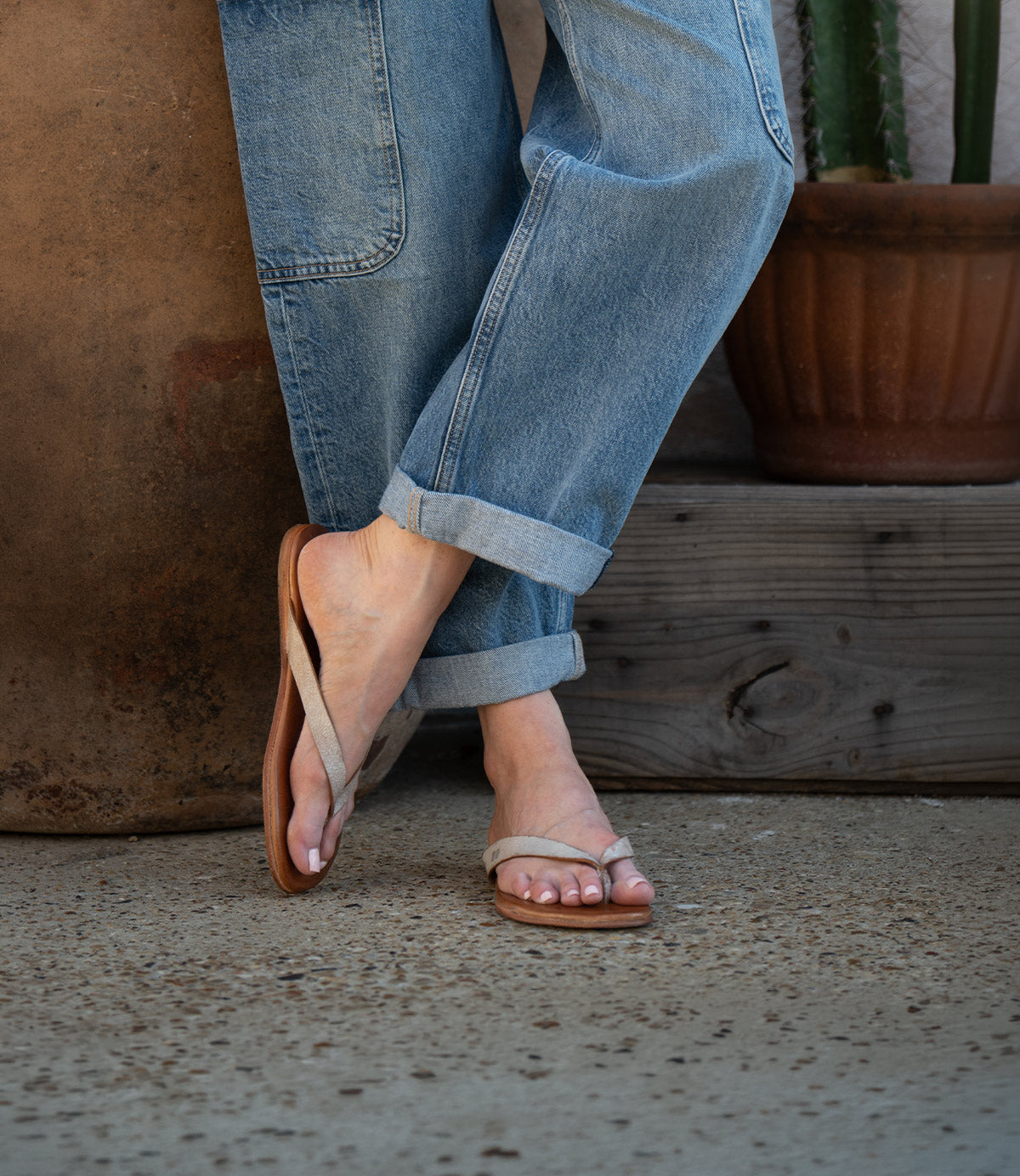 A person wearing rolled-up blue jeans and Bed Stu's Calma sandals stands on a textured concrete surface next to a potted cactus.