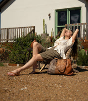 A woman wearing Bed Stu's Calma sandal relaxes on a wooden chair outside, eyes closed, arms behind her head, with a large brown bag resting beside her.