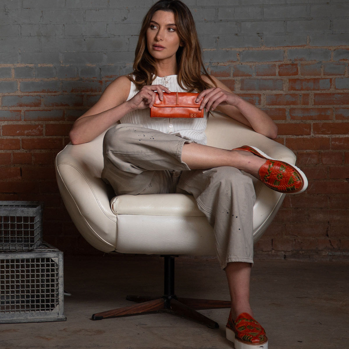 A woman sits in a white chair, holding the orange Canton 2-in-1 leather wallet by Bed Stu. She wears a white sleeveless top, beige pants, and patterned orange slip-on shoes. A metal box is nearby on the floor.