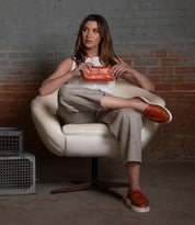 A woman sits in a white chair, holding the orange Canton 2-in-1 leather wallet by Bed Stu. She wears a white sleeveless top, beige pants, and patterned orange slip-on shoes. A metal box is nearby on the floor.