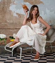 A woman in a white dress sits on a bench, smiling as she holds the versatile Bed Stu leather wallet named Canton. She wears brown shoes and is surrounded by flowers and a checkered rug against a textured, worn wall.