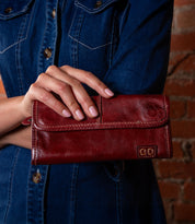 A person in a denim shirt holds the Bed Stu Canton, a stylish red leather wallet clutch, in front of a brick wall.