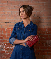 A woman in a denim dress stands before a brick wall, holding the Bed Stu Canton red leather wallet in her left hand and looking to the side.
