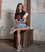 In a rustic setting, a woman sits on a wooden box, wearing the Bed Stu Canton graphic t-shirt with denim shorts and white sandals. Showcasing versatile style, she effortlessly clutches her purse and smiles while gazing slightly to the side.