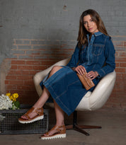 Seated on a white chair, a person wears a blue denim dress and brown sandals, holding the versatile Canton clutch by Bed Stu. Nearby, flowers rest on a metal crate with a classic brick wall backdrop.