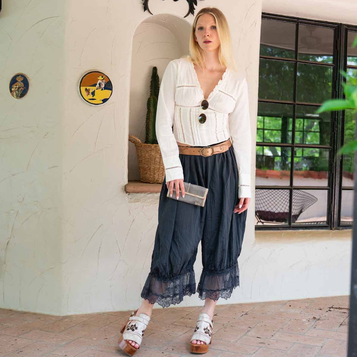 A woman stands indoors by a window, wearing a white blouse, dark culotte pants with lace trim, white sandals, and holding the Bed Stu Canton 2-in-1 wallet. Dolphin decor and plants are visible in the background.