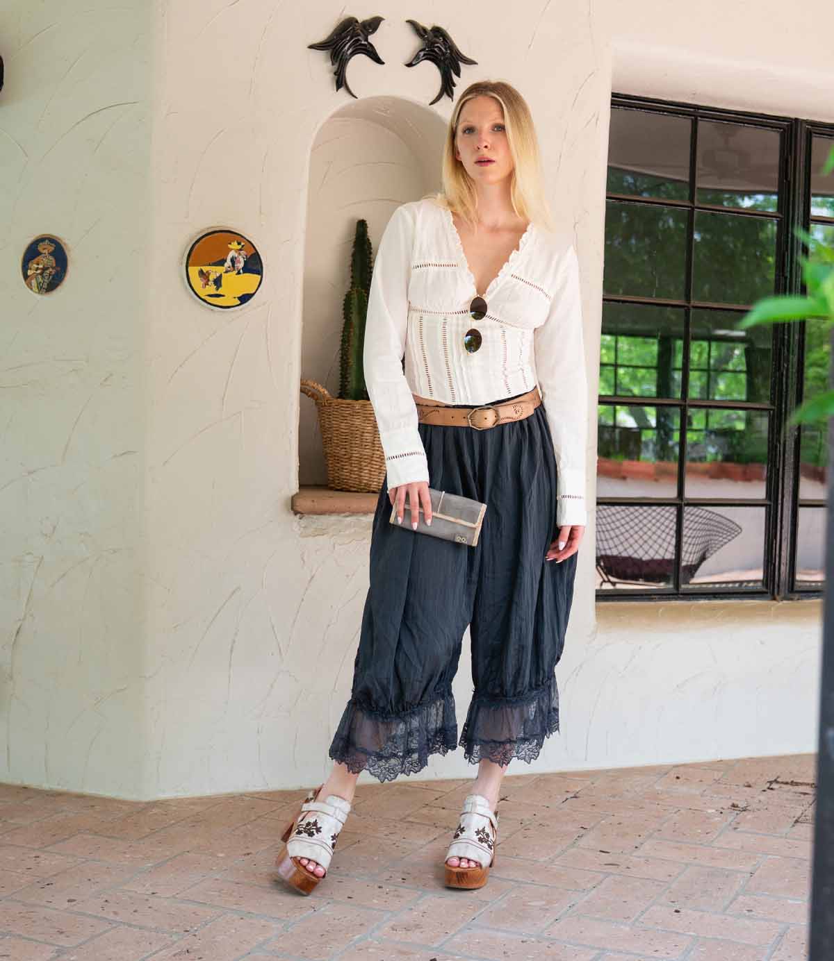 A woman stands indoors by a window, wearing a white blouse, dark culotte pants with lace trim, white sandals, and holding the Bed Stu Canton 2-in-1 wallet. Dolphin decor and plants are visible in the background.