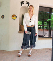 A woman stands indoors by a window, wearing a white blouse, dark culotte pants with lace trim, white sandals, and holding the Bed Stu Canton 2-in-1 wallet. Dolphin decor and plants are visible in the background.